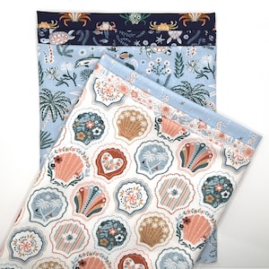 May include: Several folded fabric pieces with various ocean-themed patterns. The top fabric has crabs and turtles on a navy background. The next fabric has dolphins and fish on a light blue background. The bottom fabric has seashell designs.