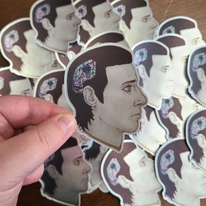 May include: A selection of stickers depicting a side profile of a person with a section of their head removed to show internal wiring and circuitry. The stickers are mainly brown, grey, and skin-toned, with colourful internal details. One sticker is held by a hand.