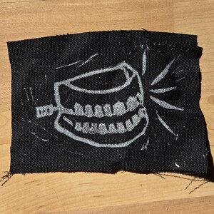 May include: A black fabric patch featuring a white illustration of a set of teeth with a toothbrush. The teeth are grinning, and radiating lines emanate from the mouth. The patch has frayed edges.
