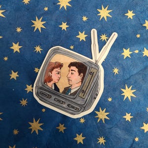 May include: A sticker of a vintage television with a couple looking at each other on the screen. The television is grey and white and the couple is drawn in a cartoon style.