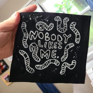 May include: Black fabric patch with white embroidered text that reads "Nobody Likes Me." The text is surrounded by white embroidered worm-like shapes.