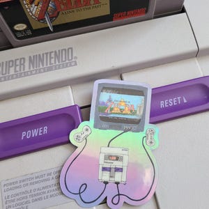 May include: A holographic sticker of a Super Nintendo Entertainment System with a controller on each side and a television screen displaying the game "The Legend of Zelda: A Link to the Past".