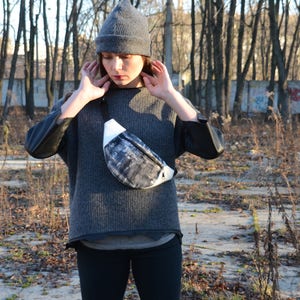 May include: A person wearing a gray beanie and a gray sweater with a black and white fanny pack.