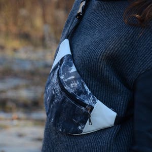 May include: A black and white patterned fanny pack with a white leather flap and a zipper closure. The fanny pack is worn across the body of a person wearing a gray sweater.
