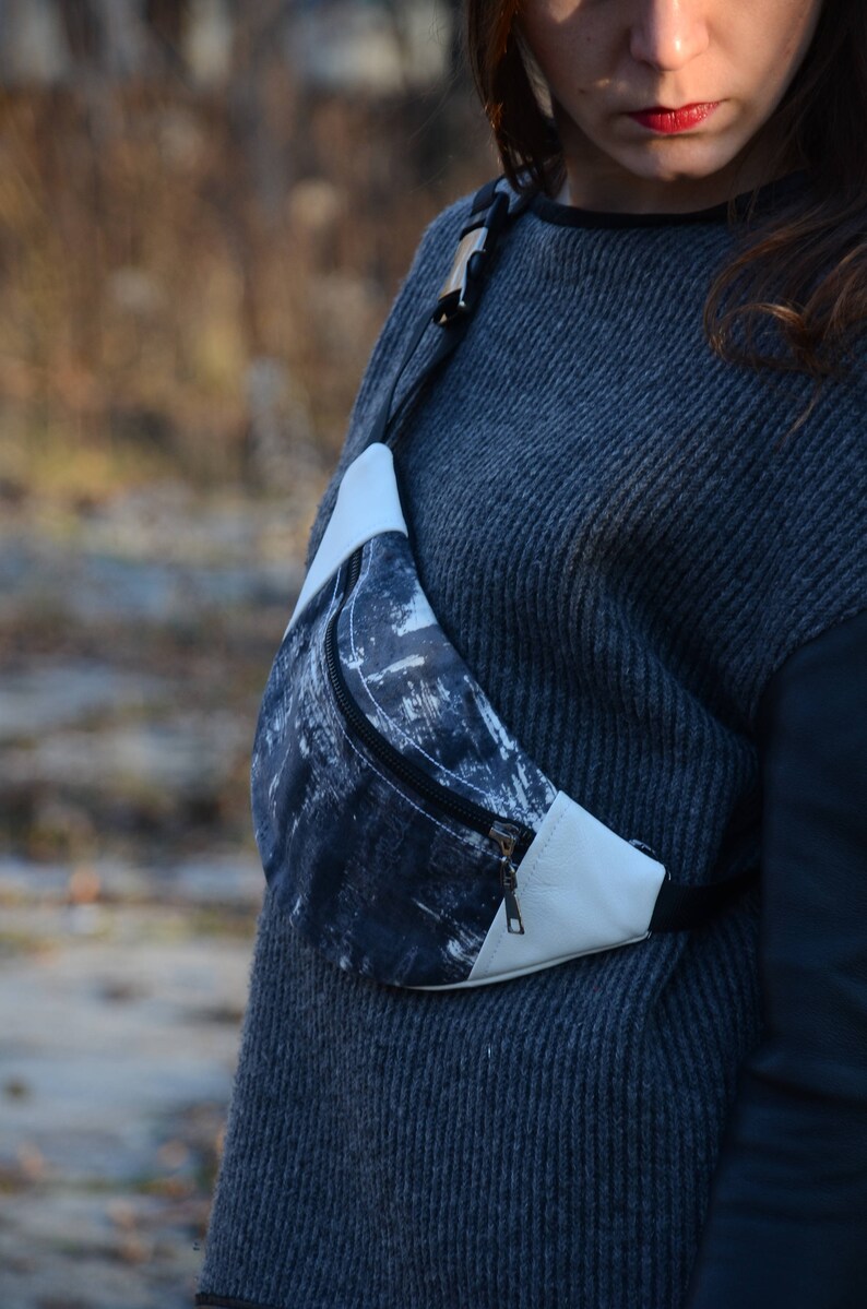 May include: A black and white patterned fanny pack with a white leather flap and a zipper closure. The fanny pack is worn across the body of a person wearing a gray sweater.