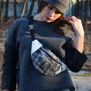 May include: A woman wearing a gray sweater with black leather sleeves and a gray beanie is wearing a black and white fanny pack with a silver buckle.