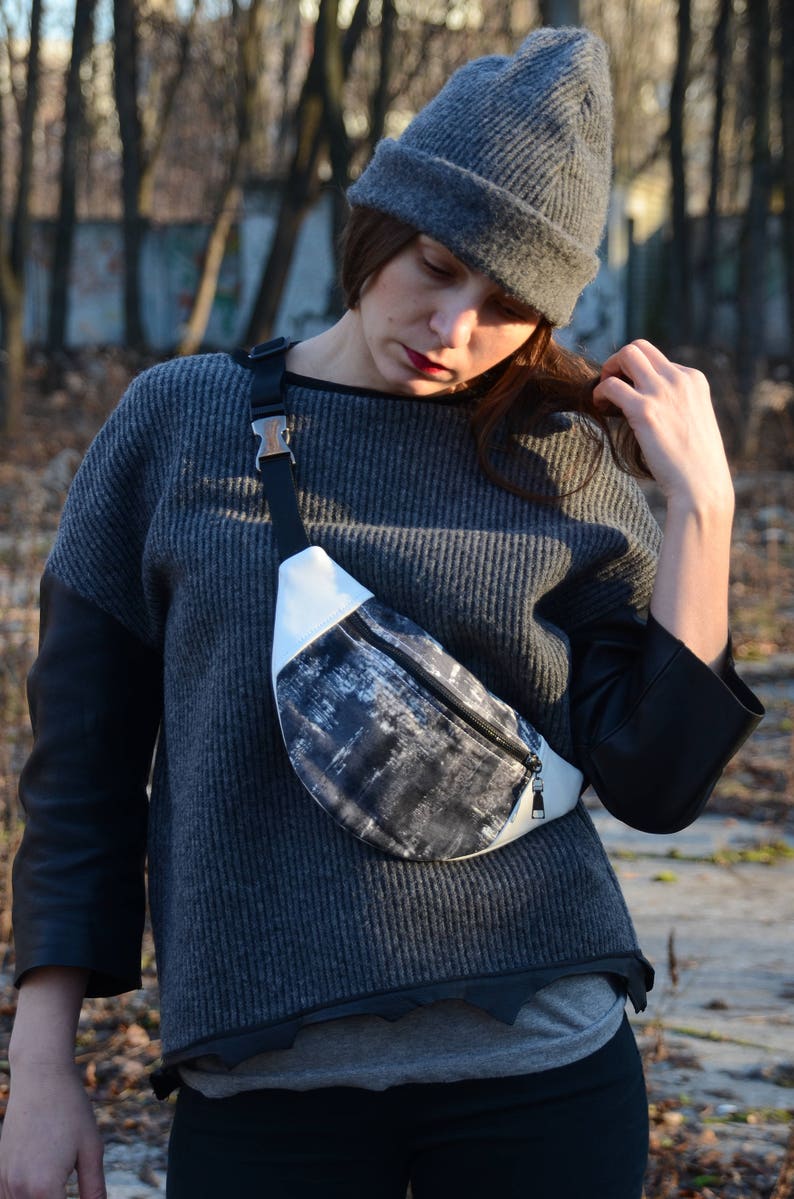 May include: A woman wearing a gray sweater with black leather sleeves and a gray beanie is wearing a black and white fanny pack with a silver buckle.