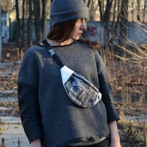 May include: A woman wearing a gray sweater with black leather sleeves and a gray beanie. She is wearing a black and white fanny pack with a silver buckle.