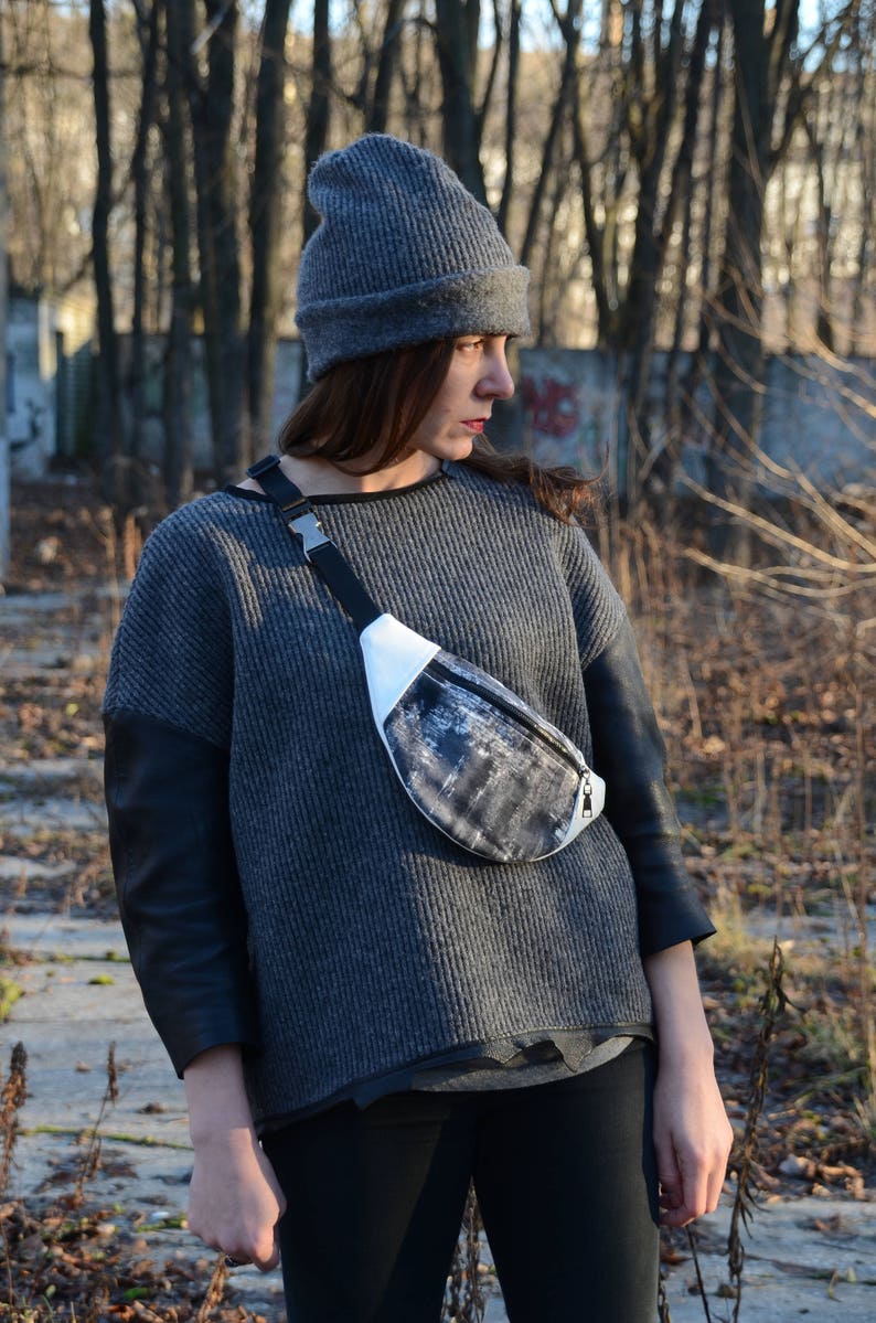 May include: A woman wearing a gray sweater with black leather sleeves and a gray beanie. She is wearing a black and white fanny pack with a silver buckle.
