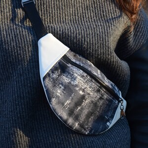 May include: A white and black patterned fanny pack with a black zipper and a black strap. The strap has a silver buckle.