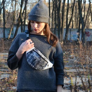 May include: A woman wearing a gray knitted beanie and a gray sweater with a black and white fanny pack.