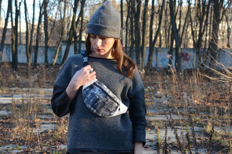 May include: A woman wearing a gray knitted beanie and a gray sweater with a black and white fanny pack.