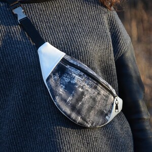 May include: A black and white patterned fanny pack with a white zipper and a silver buckle. The fanny pack is worn by a person wearing a gray sweater.