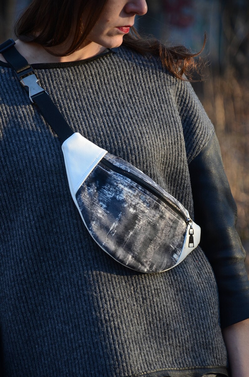 May include: A black and white patterned fanny pack with a white zipper and a silver buckle. The fanny pack is worn by a person wearing a gray sweater.