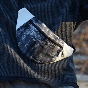 May include: A black and white patterned fanny pack with a white zipper and a white strap. The fanny pack is worn around the waist of a person wearing a gray sweater.