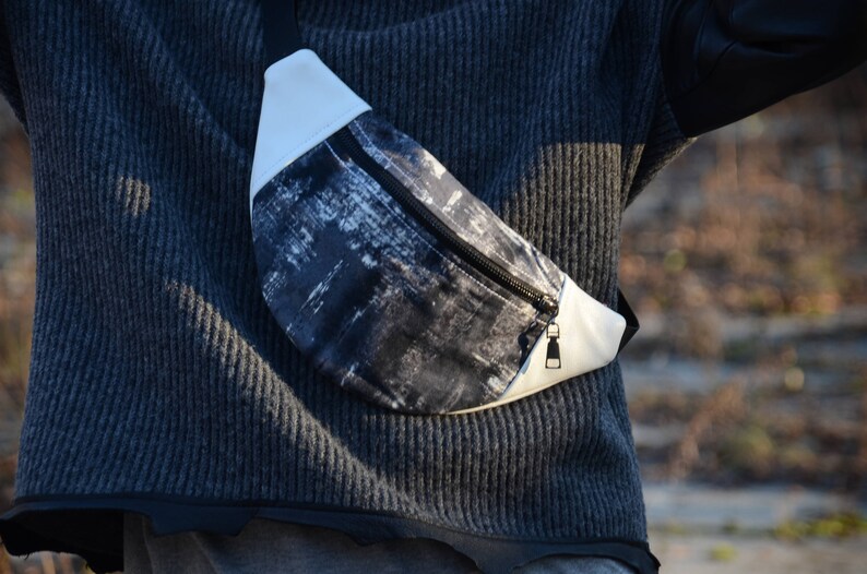 May include: A black and white patterned fanny pack with a white zipper and a white strap. The fanny pack is worn around the waist of a person wearing a gray sweater.