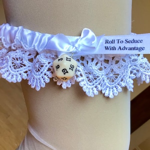 May include: White lace garter with a satin bow and a small, off-white die charm. The garter features the text "Roll To Seduce With Advantage" on a white ribbon. The garter is worn on a leg.