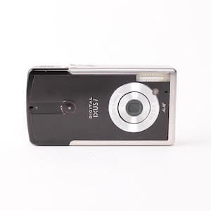 Może przedstawiać: Czarno-srebrny aparat kompaktowy Canon Digital IXUS i z centralnie umieszczonym okrągłym obiektywem, wbudowaną lampą błyskową w prawym górnym rogu oraz napisem Digital IXUS i nadrukowanym na przedniej części prostokątnego urządzenia.