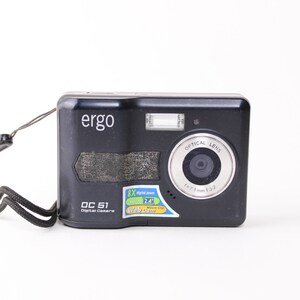 May include: A black Ergo DC 51 digital camera with a wrist strap attached to the side. The front features a lens with a silver ring, a flash, and a sticker displaying 8X digital zoom, 2.4 inch screen, and webcam functionality text.