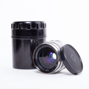May include: A vintage camera lens with a black lens cap and a black storage case. The lens is silver with black markings and a glass lens. The lens cap is off, revealing the lens. The case is cylindrical.
