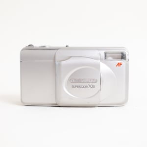 May include: A silver Olympus Superzoom 70G compact film camera with a sliding lens cover. The front features the brand name Olympus and the model name Superzoom 70G printed in grey, along with a red AF logo near the rectangular flash unit.