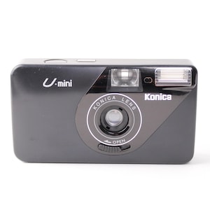 Op de afbeelding: Een zwarte Konica U-mini filmcamera. De camera heeft een flitser, een lens met de tekst "Konica Lens" en een draaiknop met "OPEN" erop. De camera heeft ook de tekst "U-mini" op de voorkant.
