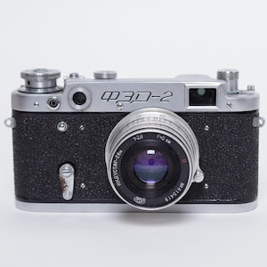 Puede incluir: Una cámara vintage FED-2 con cuerpo plateado y empuñadura negra texturizada. La lente es prominente, con el texto "ИНДУСТАР-26" visible. La cámara tiene un diseño clásico, con diales y visor. La lente tiene un tono púrpura.