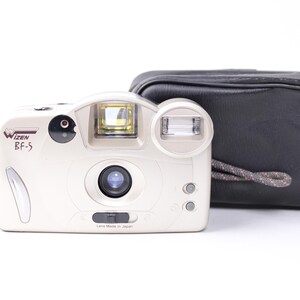Puede incluir: Una cámara Wizen BF-5 de color crema con flash y lente integrados, acompañada de una funda de transporte negra. La cámara tiene una correa para la muñeca y el texto "Lens Made in Japan".