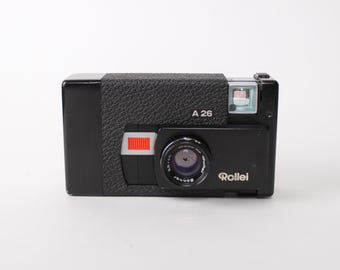 May include: Black Rollei A 26 camera with a textured grip. The camera has a lens, flash, and a red and white detail on the front. The text "A 26" is above the flash and "Rollei" is on the front.