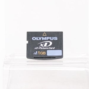 Puede incluir: Una tarjeta xD-Picture Card Olympus negra con texto blanco y una pegatina azul. La tarjeta tiene el logotipo de Olympus, el texto "xD-Picture Card" y "1GB". La tarjeta rectangular tiene una pequeña muesca en el lateral.