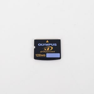 May include: A black Olympus xD-Picture Card memory card with a 128MB storage capacity. The rectangular card features a yellow triangle icon, the Olympus brand name in blue, the xD logo in yellow, and a small light blue rectangular label area.