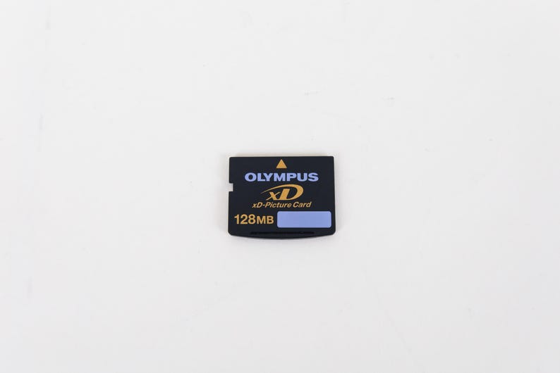 May include: A black Olympus xD-Picture Card memory card with a 128MB storage capacity. The rectangular card features a yellow triangle icon, the Olympus brand name in blue, the xD logo in yellow, and a small light blue rectangular label area.