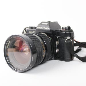 Puede incluir: Una cámara de película Pentax negra con una lente negra y una correa de cámara negra.