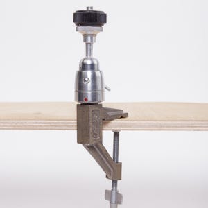 May include: A silver and black camera mount with a clamp base, attached to a wooden surface. The mount has a ball head and a screw for adjusting the camera angle. The clamp is made of metal and has a screw for attaching to a table or other surface.