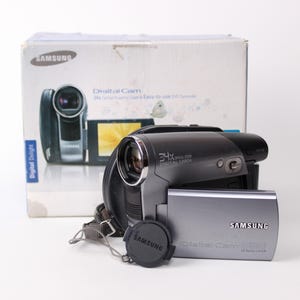 Könnte beinhalten: Schwarzer Samsung Digital-Camcorder mit Objektivdeckel und Trageriemen vor der Originalverpackung. Die Box zeigt ein Bild des Camcorders und die Worte "Digital Cam". Auf dem Camcorder steht "34x Optical Zoom".