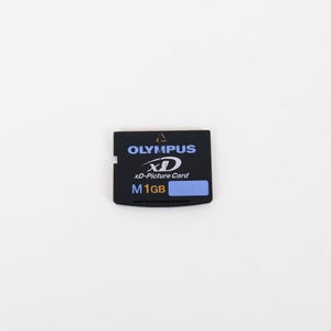 May include: A black Olympus xD-Picture Card with a 1GB storage capacity. The rectangular memory card features white text reading OLYMPUS and xD-Picture Card, with a blue label displaying the letter M and the text 1GB in white and yellow.