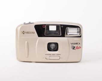 May include: A beige Yashica EZ Mate 35mm film camera. The camera has a built-in flash, a Kyocera lens, and the text "Yashica EZ Mate" in red. The camera is on a white background.