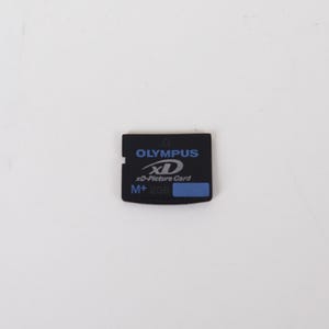 May include: A black Olympus xD-Picture Card M+ 2GB memory card. The rectangular card features blue text and branding on the front, including the Olympus logo, the xD-Picture Card name, and a small blue rectangular label on the bottom right side.