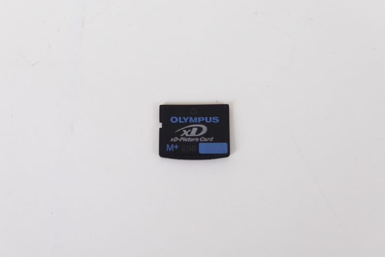 May include: A black Olympus xD-Picture Card M+ 2GB memory card. The rectangular card features blue text and branding on the front, including the Olympus logo, the xD-Picture Card name, and a small blue rectangular label on the bottom right side.