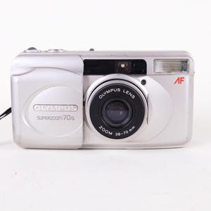 May include: A silver Olympus Superzoom 70G point-and-shoot camera with a black lens barrel. The front features the text Olympus Superzoom 70G, a black lens labeled Olympus Lens Zoom 38-70 mm, a flash unit, and a red AF logo. A black wrist strap is attached.
