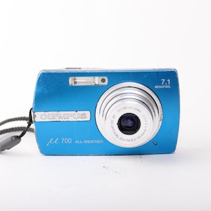 May include: A bright blue Olympus digital camera with a silver lens, featuring the text OLYMPUS, 7.1 MEGAPIXEL, and 700 ALL-WEATHER on the front. A grey wrist strap is attached to the side of the compact, rectangular camera body.