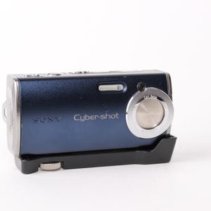 May include: A navy blue Sony Cyber-shot digital camera with a silver lens ring and a black base. The camera has the brand name and model name printed on the front. The camera is on a white background.