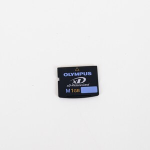 May include: A black Olympus xD-Picture Card with a 1GB storage capacity. The rectangular memory card features the Olympus brand name in white text, the xD logo, and a blue label indicating the M 1GB capacity on its front surface.