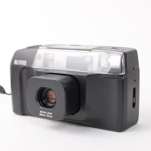 Puede incluir: Una cámara de película de 35 mm negra y plateada con flash incorporado y una correa de muñeca. La cámara tiene una lente que dice "RICOH LENS 38mm f/7.0mm".