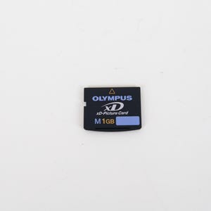 May include: A black Olympus xD-Picture Card with a 1GB storage capacity. The rectangular memory card features white and blue text, a yellow triangle icon, and a small blue rectangular label on the front surface for identification purposes.