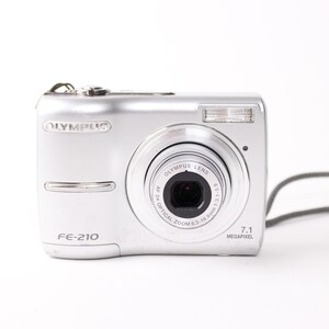 May include: Silver Olympus FE-210 digital camera with a zoom lens and a built-in flash. The camera has a 7.1 megapixel resolution and a grey wrist strap. The camera is on a white background.