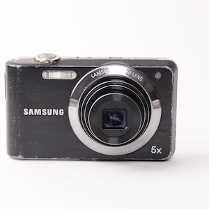 May include: A black Samsung digital camera with a silver lens and flash. The camera has the word "SAMSUNG" printed on the front. The lens has "5x" printed on the front. The camera is on a white background.