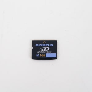 May include: A black Olympus xD-Picture Card memory card with a rectangular shape. The front label features the Olympus logo, the xD logo, and the text M 1GB in white and blue, with a small yellow triangle icon at the top center edge of the card.