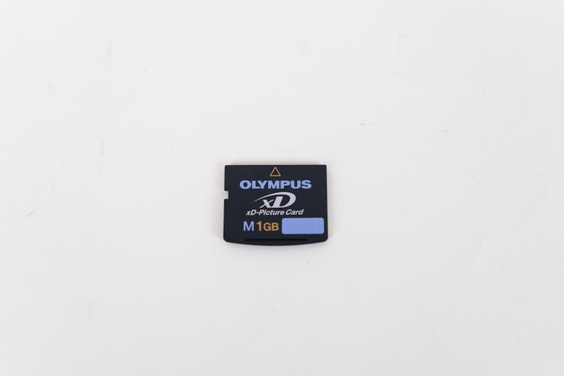 May include: A black Olympus xD-Picture Card memory card with a rectangular shape. The front label features the Olympus logo, the xD logo, and the text M 1GB in white and blue, with a small yellow triangle icon at the top center edge of the card.