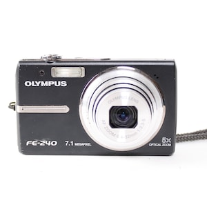 May include: A black Olympus FE-240 digital camera with a silver lens barrel. The front features the text OLYMPUS, FE-240, 7.1 MEGAPIXEL, and 5X OPTICAL ZOOM. A silver horizontal accent bar is on the left, and a gray wrist strap is attached to the side.